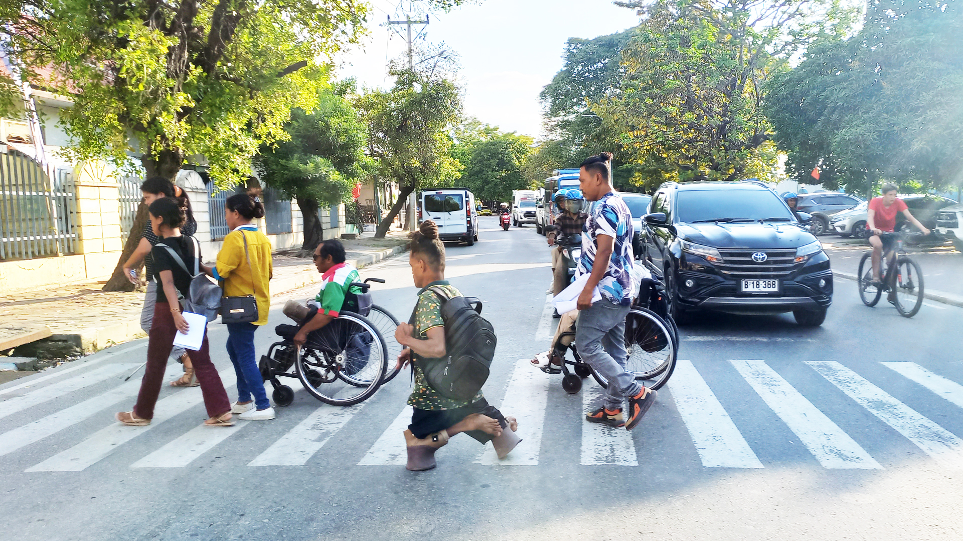 A group of people are crossing a road using a zebra crossing in Dili, Timor-Leste. Some of the people are in wheelchairs or use walking aids. Cars and bikes wait as they cross.