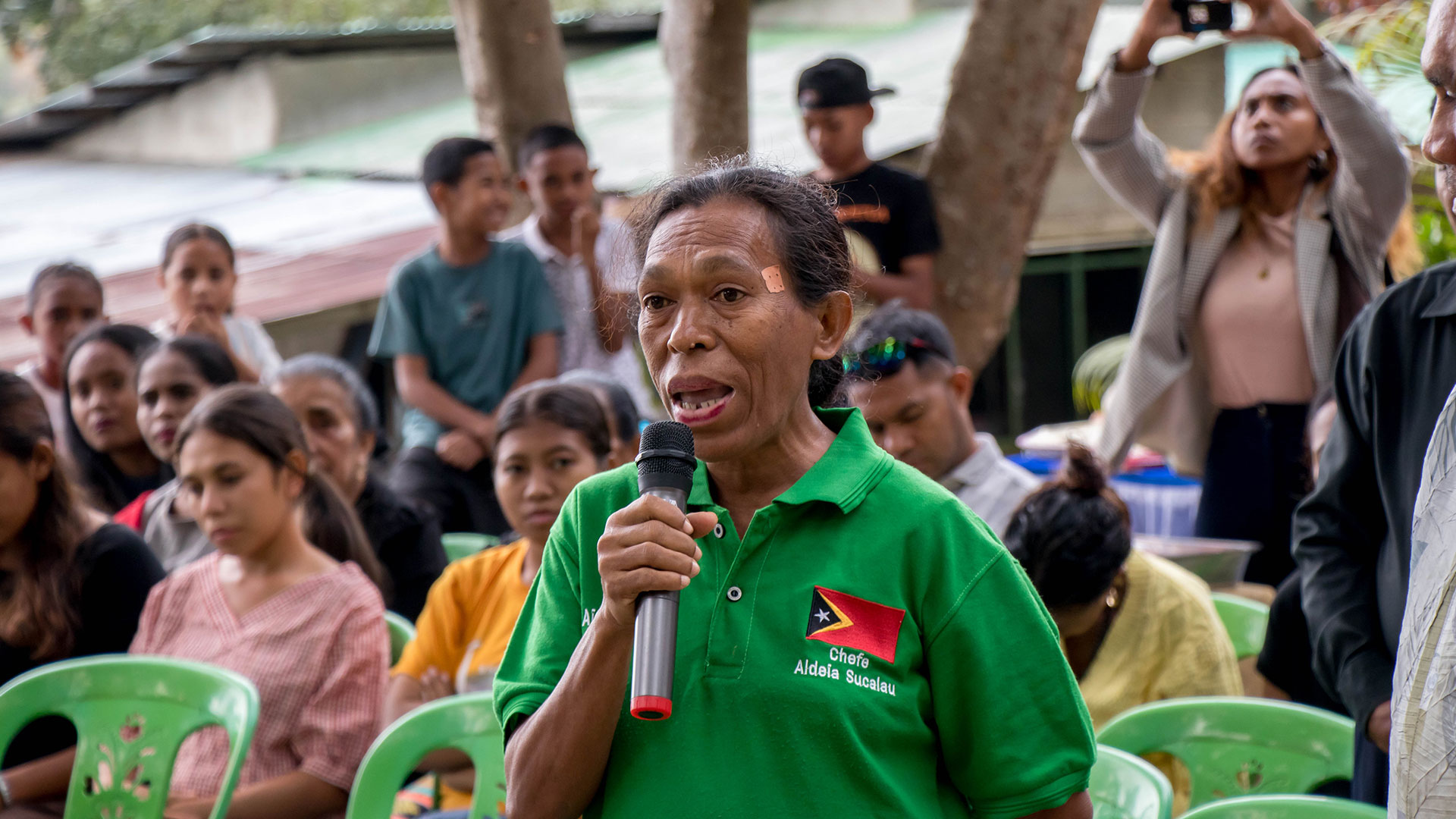 A woman in a green shirt indicating her position as the Chief of Aldeia Sucalau speaks into a microphone as she shares with delegates the benefits of the PNDS water tank, water tap and road that have been installed for the community.