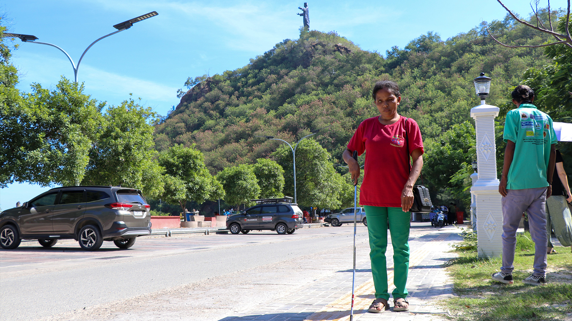 A woman with vision impairment is wearing a red top and green pants, using a mobility cane to navigate a footpath in the car park below the Cristo Rei of Dili.